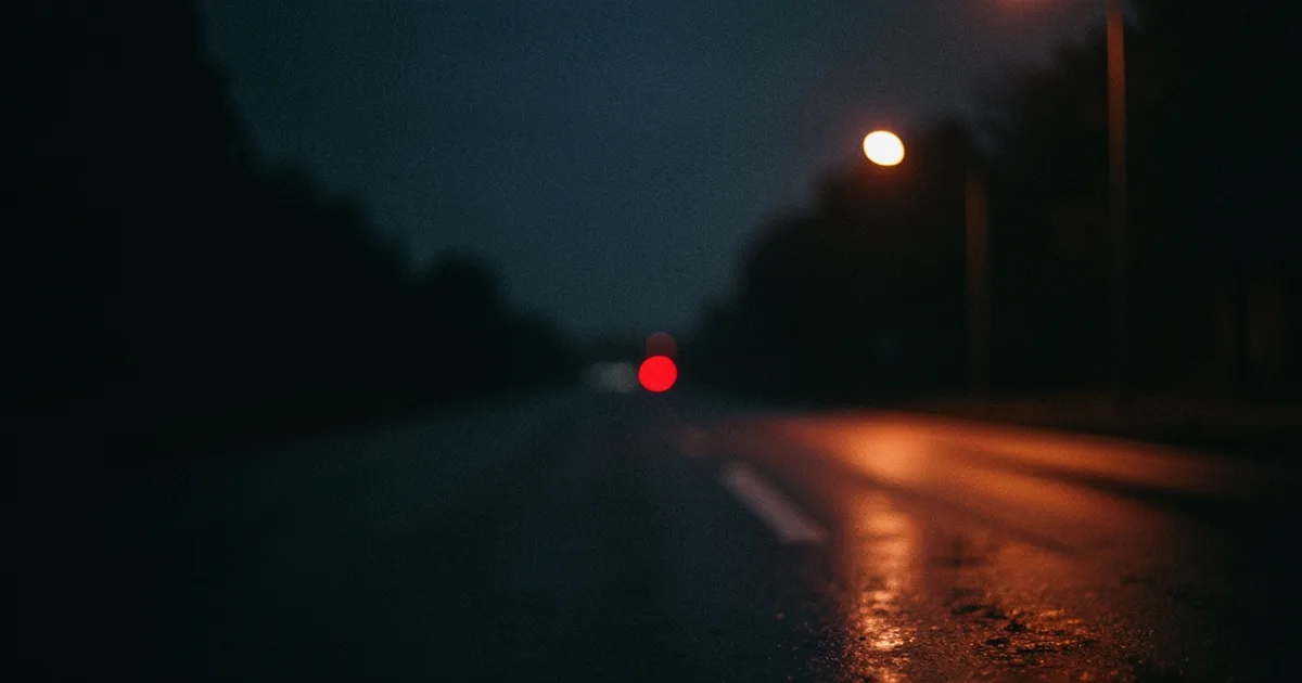 A dark wet road at night with one blurred red light in the distance
